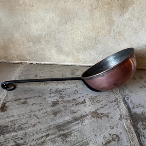 Pode incluir: Uma concha de cobre com um cabo longo e preto e um redemoinho decorativo na extremidade. A tigela da concha tem uma forma arredondada e uma cor castanho-avermelhada brilhante. O cabo é fino e reto.