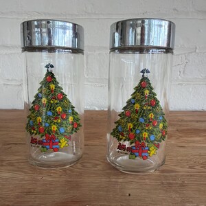 May include: Two clear glass canisters with cylindrical shapes and metallic silver lids. Each canister features a colorful graphic of a decorated Christmas tree with ornaments, a star topper, and wrapped gifts at the base, including a small toy train.