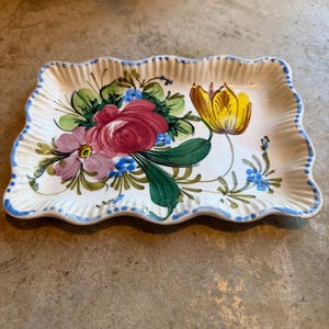 May include: A rectangular ceramic tray with a scalloped, fluted edge featuring a hand-painted floral design. The center displays a large pink rose, a smaller light purple flower, and a yellow tulip with green leaves and small blue accents.