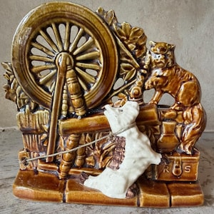 Può includere: Statuina in ceramica marrone raffigurante un filatoio, un cane e un gatto. Il cane è bianco con macchie marroni ed è attaccato alla ruota con una catena d'argento. Il gatto è marrone e siede su una piccola cassa. La statuina ha una finitura lucida.