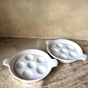 Könnte beinhalten: Zwei weiße Schneckenteller aus Keramik. Jedes Gericht hat sieben kreisförmige Vertiefungen zum Halten von Schnecken, mit einem dekorativen Rand und zwei Griffen. Die Gerichte stehen auf einer hellbraunen Oberfläche.