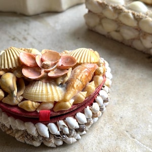 May include: A round, handcrafted seashell box with a red interior and a small red ribbon. The box is adorned with various seashells in shades of white, cream, and peach. The lid is decorated with larger, fan-shaped shells.