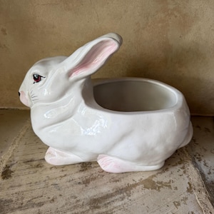 May include: A white ceramic bunny planter with pink accents on the ears and paws. The bunny is in a seated position with an open container on its back. The planter is designed for small plants or decorative items.
