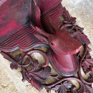 May include: A wall-mounted shelf featuring a carved deer head as the central support. The piece is finished in a distressed dark red stain and decorated with intricate, carved leaf and vine motifs surrounding the deer head and shelf base.