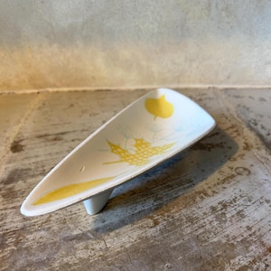 May include: A white ceramic dish with a triangular shape and a single foot. The dish is decorated with yellow leaf and floral designs. The background is a weathered wooden surface.