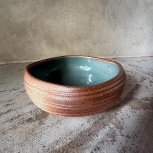 May include: A handmade ceramic bowl with a textured, reddish-brown exterior and a smooth, teal-colored interior. The bowl has a rounded shape with a slightly irregular rim. The exterior features horizontal, ridged detailing. The bowl is likely used for serving food or as a decorative piece.