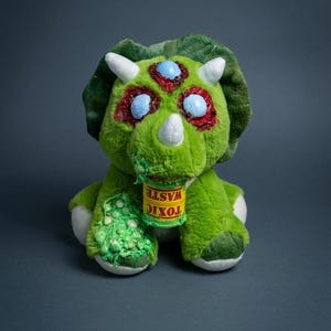 May include: A green plush triceratops toy with three eyes and white horns. The toy has red and blue accents around the eyes and a small can labeled "TOXIC WASTE" hanging from its mouth. The toy is sitting on a dark gray surface.