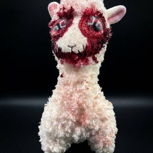 May include: A plush llama toy with white, textured fur featuring red, blood-like stains around the eyes and mouth. The toy has large, glossy, dark-centered eyes and black stitched detailing across the mouth area, creating a distressed appearance.