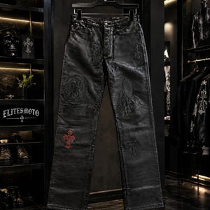 May include: Black leather pants featuring a straight-leg cut, a button fly, and multiple embossed cross motifs throughout. One prominent cross on the lower left leg is highlighted in a deep red color, contrasting with the black leather texture.