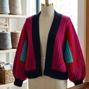 Op de afbeelding: Een felgekleurd, open vest in roze, rood en blauwgroen, gepresenteerd op een mannequin. De trui heeft een colorblock-ontwerp met marineblauwe afwerking en een getextureerde, dikke brei. De mouwen zijn lang en volumineus.