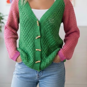 May include: A green and pink cardigan with a V-neck and wooden buttons. The sleeves are pink, and the body is green with a textured pattern. The cardigan is paired with blue jeans.