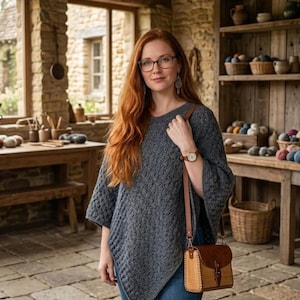 May include: A woman wearing a gray crocheted poncho and blue jeans, with a brown leather purse. The background shows a rustic room with stone walls, wooden shelves, and baskets filled with yarn. The woman is wearing glasses and a watch.