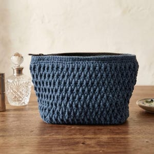 May include: A blue crochet cosmetic bag with a black zipper, sitting on a wooden surface. The bag has a textured, woven appearance. Lipstick tubes, a perfume bottle, and a small dish are in the background.