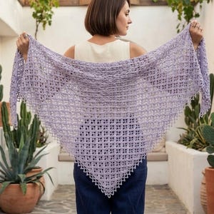 Puede incluir: Un chal de ganchillo lila claro, de tejido abierto y forma triangular. El chal se muestra abierto, mostrando su delicado patrón y borde festoneado. El fondo incluye plantas y una alfombra estampada.