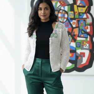 May include: A white, open-weave crochet cardigan with floral embellishments. The cardigan is paired with a black top and emerald green pants. The background features a colorful abstract painting.