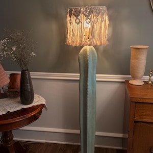 May include: A floor lamp featuring a tall, light green, vertical cactus-shaped base and a cylindrical lampshade covered in cream-colored macrame cord with intricate knotted patterns and long, hanging fringe that glows when illuminated.