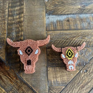 May include: Two decorative bull skull ornaments. The skulls are a copper color and covered in small beads. Each skull has horns and a geometric design in the center. One has a black and white design, and the other has a yellow and black design.