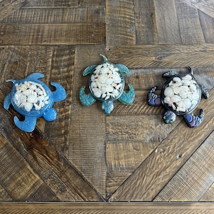Peut inclure: Trois figurines décoratives de tortues. Chaque tortue est remplie de petits coquillages blancs. Les tortues sont dans des tons de bleu, de sarcelle et de noir. Les figurines sont exposées sur une surface en bois.