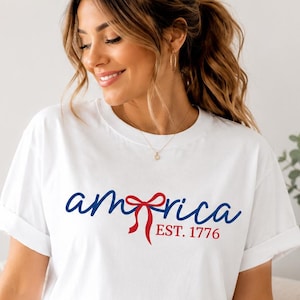 Op de afbeelding: Een wit T-shirt met korte mouwen voorzien van de blauwe tekst "america" waarbij een rode strik de letter "e" vervangt, met daaronder de rode tekst "EST. 1776". Het shirt heeft een ronde hals en opgerolde mouwen voor een strakke uitstraling.