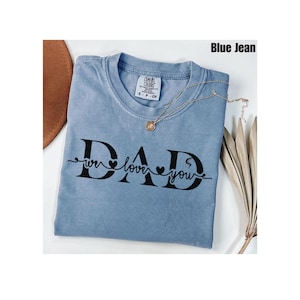 May include: A blue jean-colored t-shirt with the words "DAD we love you" in black script. The shirt has a crew neck and short sleeves. A gold necklace is draped over the shirt. The shirt is folded on a white surface.