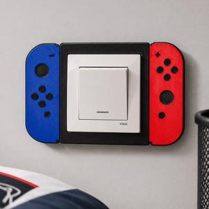 Op de afbeelding: Een witte Viko-lichtschakelaar aan een muur, omlijst door een zwarte kunststof behuizing die lijkt op een gamecontroller met een blauwe linkerkant en een rode rechterkant, voorzien van zwarte ronde knoppen en richtingsknoppen.