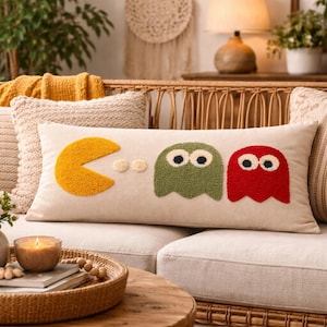 Puede incluir: Un cojín rectangular beige con un diseño de Pac-Man y fantasmas. El Pac-Man es amarillo y los fantasmas son verdes y rojos. El cojín está sobre un sofá crema con otros cojines decorativos.