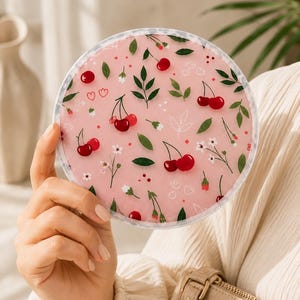 May include: A round compact mirror featuring a light pink background decorated with a repeating pattern of red cherries, green leaves, and small white flowers. The circular frame is clear, and the surface displays a smooth, glossy finish.