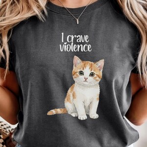 Puede incluir: Una camiseta gris carbón de cuello redondo con un gráfico de un gatito atigrado naranja y blanco sentado. Sobre el gatito, el texto "I crave violence" aparece en una fuente blanca estilizada. La camiseta tiene mangas cortas y corte estándar.