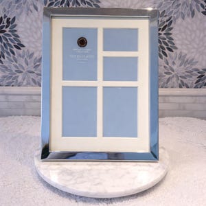 May include: A silver-plated multi-photo frame with a white border and light blue backing. The frame has four openings and is displayed on a white marble stand. The text "MARTHA STEWART COLLECTION SILVER PLATED" is visible.