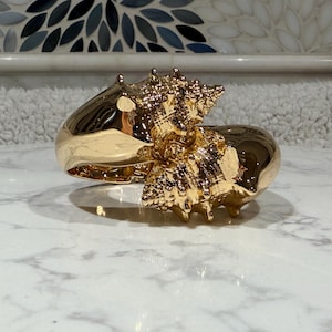 May include: A polished gold-tone hinged cuff bracelet featuring a prominent, textured seashell design. The metallic surface is highly reflective, showcasing the intricate spiral and spiked details of the shell motif across the front of the piece.