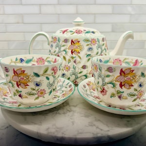 May include: A white porcelain tea set with floral patterns. The set includes a teapot, two teacups, and saucers. The floral design features pink, yellow, and blue flowers with green accents. The rims of the cups and saucers have a green trim.