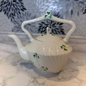 May include: Cream-colored ceramic teapot with a curved handle and a lid. The teapot is decorated with green shamrock designs. The handle and lid are accented with green shamrock details. The teapot is on a white surface.