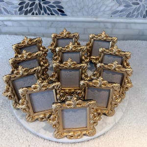 May include: A collection of ornate, gold-colored picture frames arranged on a white, round surface. The frames have a detailed, vintage design with decorative flourishes. The frames are empty, ready for photos.