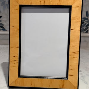May include: A rectangular picture frame with a light-colored wood grain border and a black inner frame. The frame is standing upright, showcasing a white interior space for a photo or artwork.