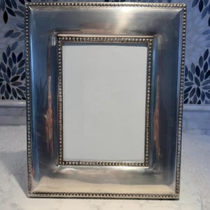May include: A rectangular silver picture frame with a beaded border. The frame has a polished finish and a white interior, suitable for displaying a photograph or artwork. The frame is designed to stand on a flat surface.
