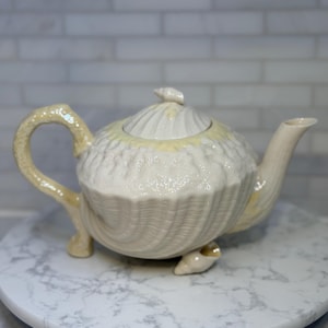 May include: Cream-colored ceramic teapot with a seashell design. The teapot has a curved handle, a spout, and a lid with a small shell-shaped knob. The teapot sits on three small feet and is displayed on a marble-patterned surface.