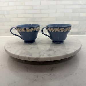 May include: Two blue teacups with gold floral trim sit on a white marble lazy susan. The teacups have a classic design with curved handles. The background is a white brick-patterned wall.