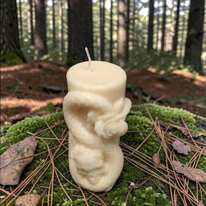 May include: Ivory-colored pillar candle with a carved dragon design. The candle features a skull and rose motif, set against a backdrop of a mossy surface and a forest. The candle is approximately 4 inches tall.
