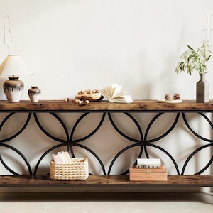 May include: A rectangular console table featuring a dark wood grain top and bottom shelf supported by a black metal frame with a repeating circular pattern. The table holds a lamp, a small vase, a bowl of nuts, an open book, and a plant.