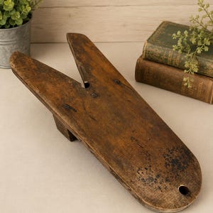 May include: A weathered brown wooden boot jack with a V-shaped notch at one end and a small circular hole at the rounded base. The surface shows natural wood grain, dark stains, and signs of age, featuring a sturdy block support underneath.