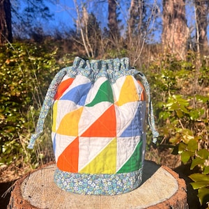 May include: A handmade drawstring bag with a patchwork design. The bag features a colorful pattern of triangles in orange, yellow, green, and blue, set against a white background. The top and base are trimmed with a floral fabric. The bag is sitting on a tree stump.