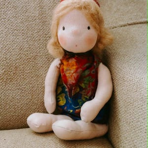 Puede incluir: Muñeca de tela hecha a mano con tez clara, cabello rubio y mejillas rosadas. La muñeca lleva una bufanda colorida y un vestido azul. Está sentada sobre una superficie tapizada beige.