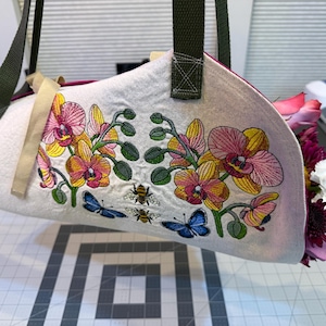 May include: A white tote bag with a floral embroidery design featuring pink, yellow, and orange orchids, green leaves, blue butterflies, and bees. The bag has green straps and is filled with colorful flowers.