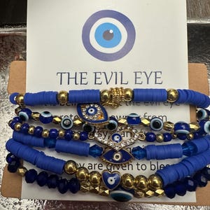 May include: Stack of blue and gold beaded bracelets with evil eye charms. The bracelets are displayed in front of a card with a blue eye graphic and the text "THE EVIL EYE". The bracelets have various bead sizes and shapes.
