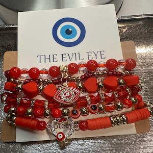 May include: Stack of red beaded bracelets with gold accents and an Evil Eye charm. The bracelets feature various bead shapes, including hearts and round beads. A card behind the bracelets reads "THE EVIL EYE" with an eye symbol.