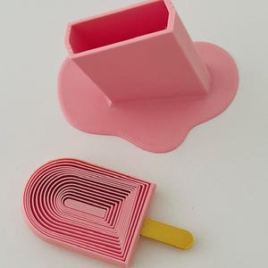 May include: A pink rectangular container with a melted-look base and a separate pink arched cookie cutter featuring a yellow handle. The cutter has a series of concentric U-shaped ridges, while the container has a hollow, open top.
