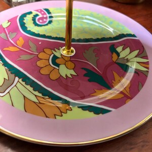 May include: A two-tiered serving tray with a gold central support. The top tier has a pink border and a decorative floral design in pink, green, yellow, and orange. The bottom tier has a pink border with a gold rim.