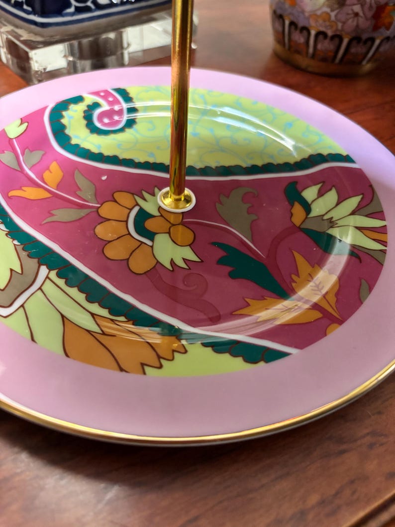 May include: A two-tiered serving tray with a gold central support. The top tier has a pink border and a decorative floral design in pink, green, yellow, and orange. The bottom tier has a pink border with a gold rim.