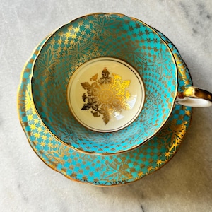 May include: A vintage teacup and saucer set. The cup and saucer are turquoise with a gold floral pattern. The cup has a gold handle and a gold rim. The inside of the cup is cream-colored with a gold floral design.