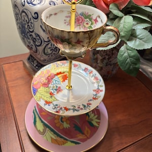 May include: A three-tiered serving stand with a gold handle. The top tier holds a floral teacup. The middle tier is a small plate with a floral design, and the bottom tier is a pink plate with a patterned design. The stand is on a wooden surface.